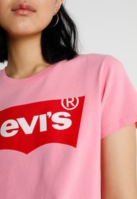 Růžové bavlněné tričko s výrazným červeným logem a bílým textem "Levi's." Hladký kulatý výstřih a krátké rukávy. Hladká textura.