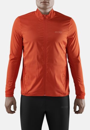 Hombre vistiendo una chaqueta deportiva de manga larga color naranja brillante con cremallera y leggings deportivos negros sobre un fondo liso.