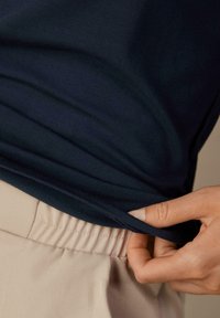 Dunkelblauses, tailliertes Hemd aus weichem Stoff, in hellbeige Hosen mit elastischem Bund gesteckt, mit einer Hand, die den Saum des Hemdes greift.