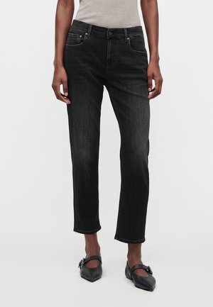 Femme portant un jean noir droit, un haut beige sans manches et des ballerines noires à bouts pointus avec des brides à boucle, debout devant un fond blanc.