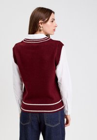 Pull sans manches bordeaux avec une finition texturée, ornements blancs rayés le long de l'encolure et de l'ourlet, associé à une chemise blanche à col.