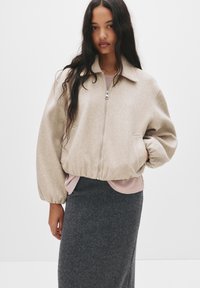 Béžova volna mešana bomber jakna z zadrgo, s poševnim ovratnikom in elastičnimi manšetami. Nosi se čez svetlo roza top in temno sivo krilo.