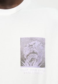 Αφηρημένο γραφικό σε μοβ και γκρι αποχρώσεις, με στριφτό, κυματιστό σχήμα πάνω από το κείμενο «Collide · Endure · Emerge A Work in Progress» σε ένα λευκό μπλουζάκι.