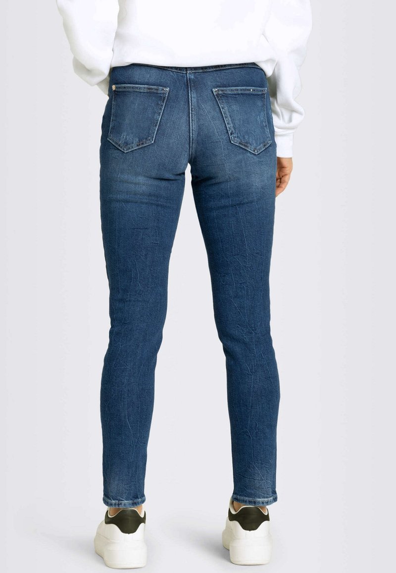 Slim-fit modré džíny s texturovaným povrchem, obsahující dvě zadní kapsy a mírné vyblednutí. Nosené s bílými a zelenými teniskami.