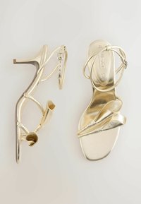 Sandale metalice aurii cu toc înalt, curele la gleznă și accente decorative răsucite în formă de petale în partea frontală.