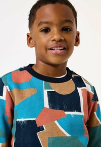 Ragazzo giovane sorridente con capelli corti che indossa una maglietta a motivo geometrico colorato con forme blu, arancione, beige e nere su sfondo bianco.