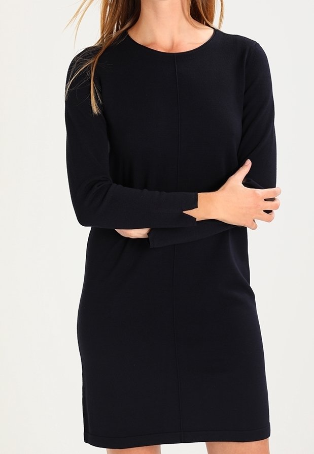 Femme portant une robe noire à manches longues, longueur genoux, avec un col rond, debout les bras croisés devant un fond clair uni.