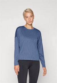 Maglietta atletica a maniche lunghe di colore blu con tessuto testurizzato, scollo rotondo e vestibilità rilassata, abbinata a leggings neri.