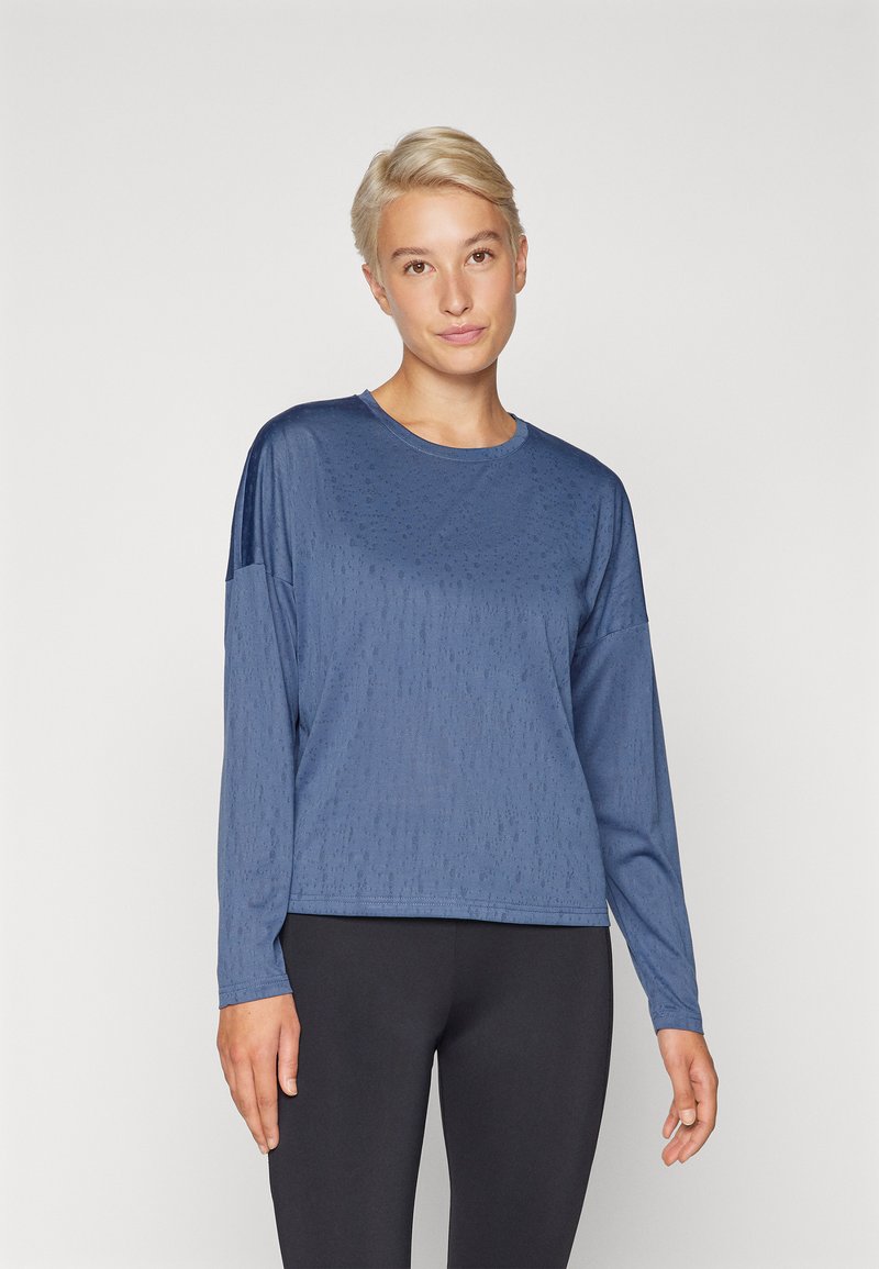 Maglietta atletica a maniche lunghe di colore blu con tessuto testurizzato, scollo rotondo e vestibilità rilassata, abbinata a leggings neri.