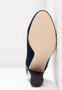 Scarpa con tacco alto in camoscio blu navy, dalla forma curva, suola liscia e tacco in gomma texturizzata. Presenta un'integrazione senza soluzione di continuità dei materiali.
