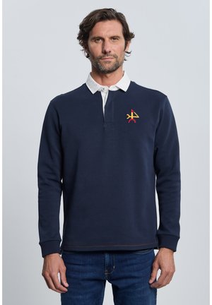 Hombre con cabello corto y oscuro y barba, vistiendo una camisa polo azul marino de manga larga con cuello blanco y vaqueros, de pie frente a un fondo liso.