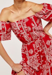 Robe rouge décolletée avec motifs floraux et géométriques blancs, taille cintrée avec un lien, et manches amples. Tissu léger.