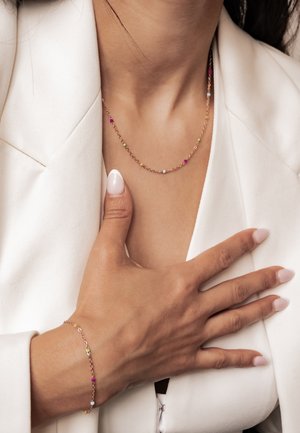 Nahaufnahme einer Person, die einen weißen Blazer trägt, mit einer filigranen goldenen Halskette und einem passenden Armband mit kleinen bunten Perlen.