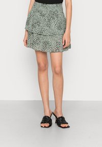 Femme portant une mini-jupe verte à pois noirs et volants, associée à des sandales à talons tressées noires sur un fond uni.