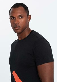 Musta puuvillapaita, jossa on pyöreä kaula-aukko ja lyhyet hihat, etupuolella on rohkea oranssi abstrakti kuvio. Sileä pinta.