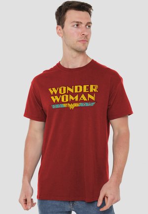 Young man wearing a red Wonder Woman T-shirt, looking to the side, standing against a plain light gray background.