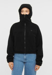 Giacca in pile nera con colletto alto e zip, caratterizzata da maniche lunghe e design corto. Morbida al tatto con un marchio ricamato sul petto.