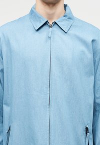 Άνδρας που φοράει ανοιχτό μπλε μπουφάν με φερμουάρ και γιακά, εμφανές φερμουάρ και τσέπες με φερμουάρ στο πλάι, σε απλό ανοιχτό φόντο.