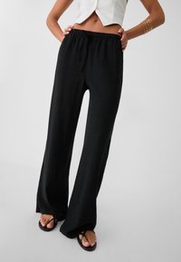 Zwarte wijde broek met een trekkoord in de taille, zachte stof en zijslits. Gecombineerd met zwarte slippers.