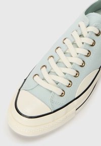 Light blue canvas sneaker with white laces, gold eyelets, and a cream rubber toe cap; black edging along the sole for contrast.