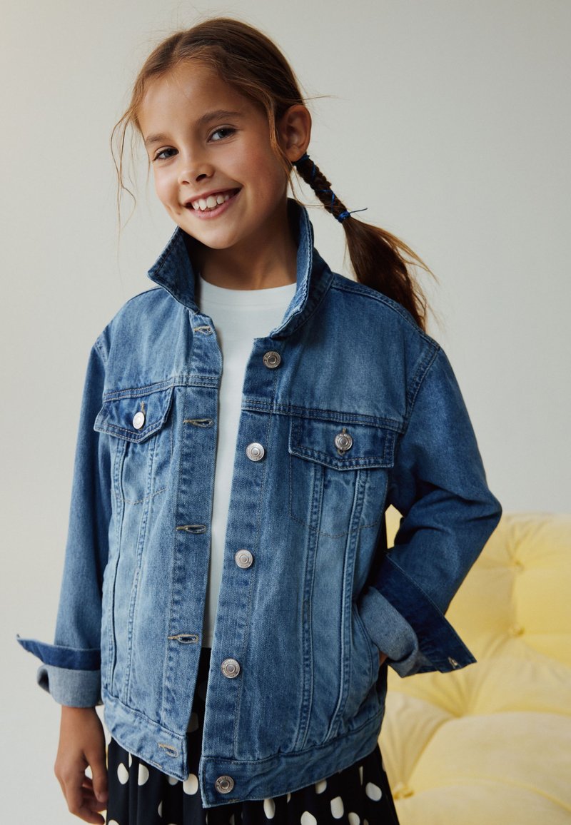 Ragazza sorridente con capelli intrecciati indossa una giacca di jeans blu sopra una camicia bianca e una gonna nera a pois bianchi, in piedi accanto a un divano giallo.