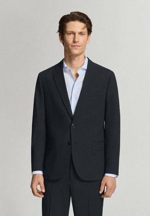Hombre con cabello corto y castaño, vestido con una chaqueta de traje oscura y camisa azul claro, de pie frente a un fondo claro y liso.