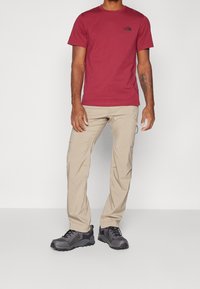 Mörk röd t-shirt med en liten svart logotyp på bröstet, ljusbeiga byxor och svarta vandringsskor med grå detaljer och texturerad sula.