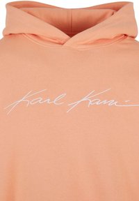 Sudadera de color melocotón hecha de un tejido suave, con un gran gráfico de firma blanca en el pecho. Presenta un diseño clásico con capucha.