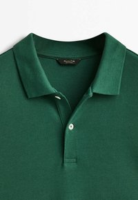 Polo shirt vert foncé, tissu texturé, col plat, patte de boutonnage à deux boutons avec boutons blancs, étiquette visible à l'intérieur du col.
