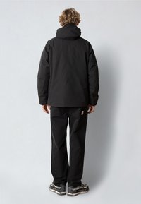 Svart vattentät jacka med huva, kraftig textur och avslappnad passform. I kombination med svarta byxor med logotypmärke och grå sneakers.