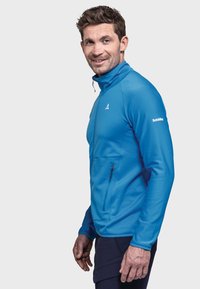 Helles blaues Zip-Jacke mit strukturiertem Muster, hohem Kragen und Reißverschluss-Tasche auf der Vorderseite. Verfügt über ein Logo am linken Arm.