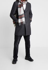 Grå uldfrakke med en skræddersyet design, hvid og rutet scarf, sorte jeans og sorte sneakers. Har knapper og lommedetaljer.