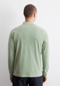 Dlhý rukáv polo tričko z mäkkej svetlozelenej bavlny s golierom a spodným lemom, ktorý sa jemne zakrivuje. Minimalistický dizajn, žiadne viditeľné vzory.