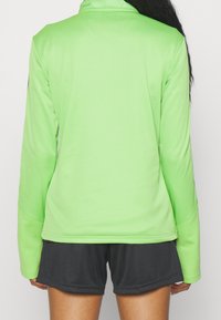 Jasnozielona sportowa kurtka z wysokim kołnierzem i długimi rękawami, zestawiona z ciemnoszarymi szortami; gładki, elastyczny materiał o dopasowanym kroju.