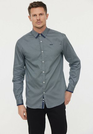 Chemise à boutons dans un bleu foncé avec un motif géométrique blanc. Elle présente un col contrasté bleu marine et des accents de boutons, des manches longues et une coupe ajustée.