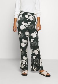 Spodnie z dzwonami wykonane z ciemnozielonego materiału w białe kwiatowe wzory. Elastyczny pasek w talii. Modelka nosi czarne i beżowe sandały.