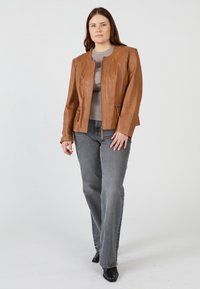 Braune Lederjacke mit Reißverschluss, Schulternähten und Seitentaschen, kombiniert mit einem grauen Top und Jeans. Die Umgebung ist schlicht und neutral.