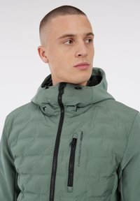 Junger Mann mit kurzem Haar, der eine grüne gesteppte Jacke mit Kapuze, einem durchgehenden Reißverschluss und einer vertikalen Brusttasche trägt, die mit "Protest High Performance" beschriftet ist.