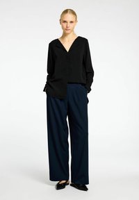 Sort langærmet bluse med V-hals og knaplukning, parret med brede navy bukser og sorte slip-on sko. Enkel og moderne design.