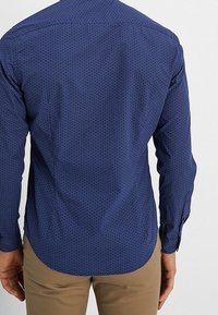 Marinblå långärmad skjorta med ett geometriskt vitt mönster, med en böjd fåll och knappslutna manschetter, tillverkad av ett mjukt tyg.