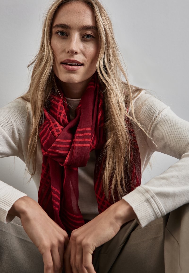 Écharpe rouge à motif rayé, fabriquée en tissu léger, drapée autour du cou, assortie à un pull crème.