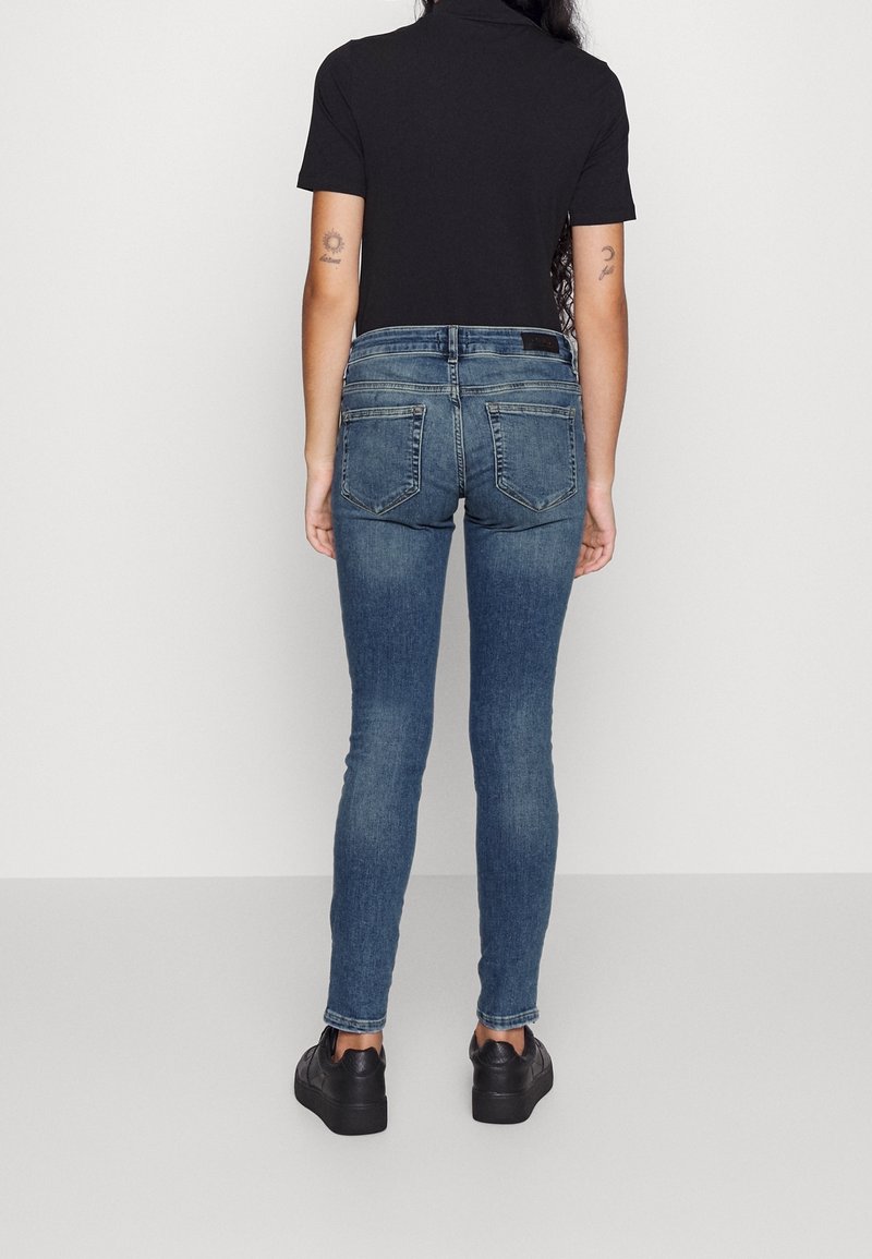 Person steht mit dem Rücken zur Kamera, trägt ein schwarzes kurzärmeliges Oberteil, blaue Skinny-Jeans mit Gesäßtaschen und schwarze Sneakers vor hellem Hintergrund.