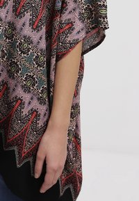 Flerfärgad kimono-inspirerad topp i lätt tyg, med djärva blommönster och geometriska mönster i röd, grön och svart mot en ljuslila bakgrund.