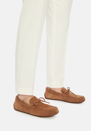 Mocassini in suede marrone con punta arrotondata, cuciture decorative e un fiocco come dettaglio. Indossati con pantaloni bianchi aderenti.
