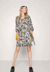 Vestido floral com mangas três quartos, decote em V e cintura justa. O padrão apresenta flores brilhantes sobre um fundo escuro. Combinado com uma bolsa amarela.