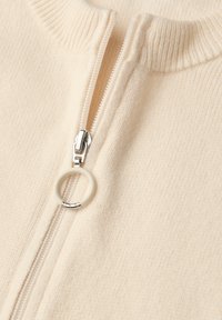 Kermanvärinen neulepusero, jossa on etutaskussa hopeinen vetoketju, pyöreä valkoinen koriste, teksturoitu kangas ja resorinen kaulus.