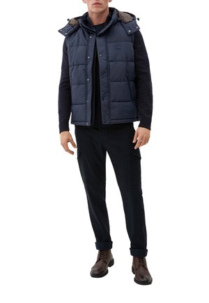 Mann in einer marineblauen gesteppte ärmellose Jacke über einem schwarzen Langarmpullover, schwarzen Cargo-Hosen und braunen Schnürboots.