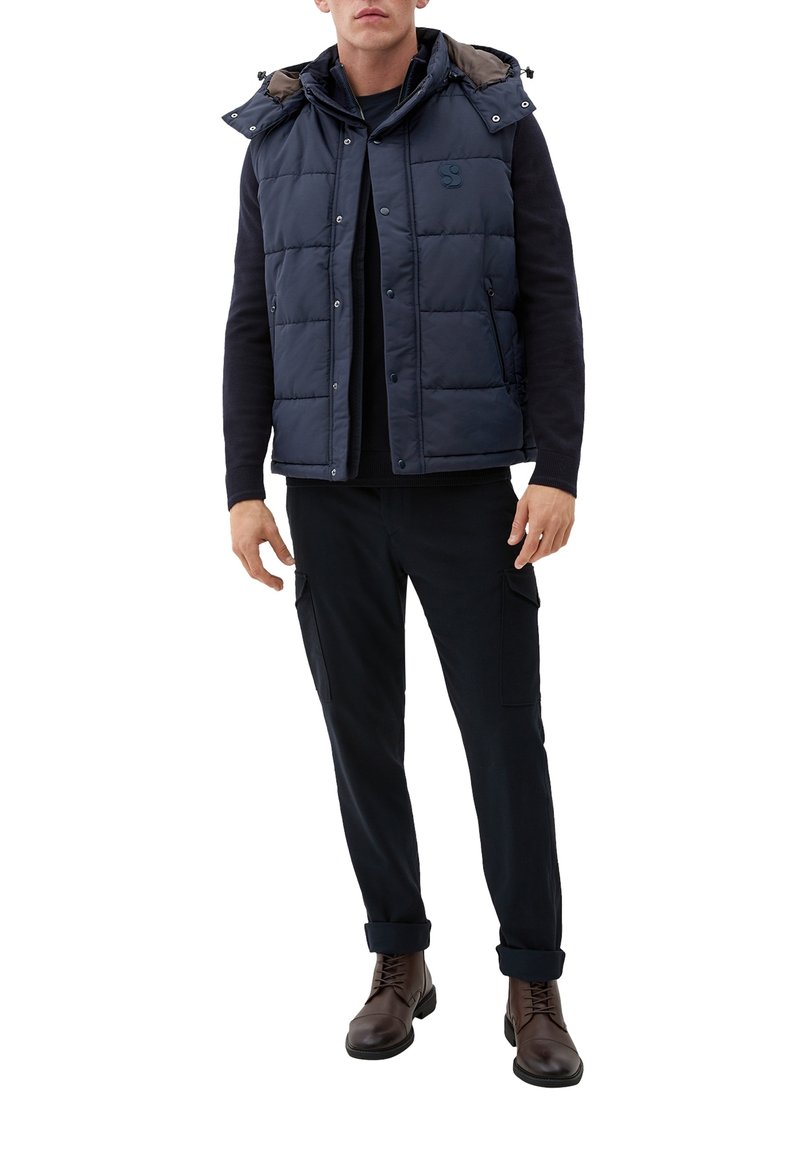 Mees, kellel on seljas tumesinine tikitud ilma käevarrта jakki, must püsivaba sviiter, mustad cargo-püksid ja pruuni nööritavad saapad.