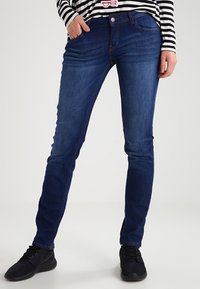 Tmavě modré slim-fit džíny s pětipocketovým designem, s jemným šitím a hladkou denimovou texturou, v kombinaci s černými teniskami.