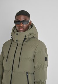 Giacca imbottita verde oliva con accessori neri, colletto alto, stringhe regolabili e tasche con zip. Tessuto testurizzato con un aspetto trapuntato.